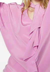 Blusa in satin rosa con design drappeggiato, dotata di un nodo al collo e vestibilità morbida. Texture morbida e finitura liscia e lucida.