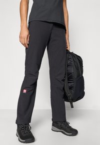 Calças de exterior pretas com uma textura suave e costuras reforçadas, apresentando um logótipo vermelho, acompanhadas de uma mochila escura e botas de caminhada pretas.