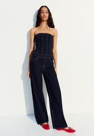 Žena stojaca, oblečená v tmavomodrý denimový overal bez ramienok a červené ploché topánky na bielom pozadí.