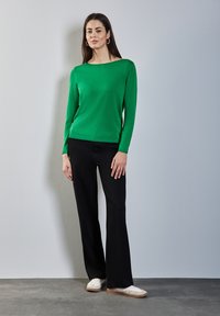 Pull vert à manches longues avec un large col, associé à un pantalon noir à jambes larges et des baskets blanches sur un sol gris texturé.