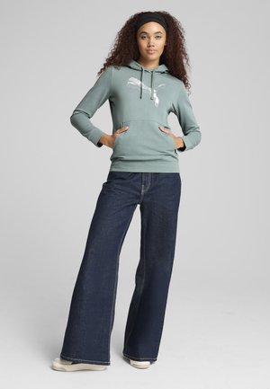 Felpa verde con tasca frontale e logo argentato, abbinata a jeans in denim scuri a gamba larga. Scarpe chiare con suole texturizzate.