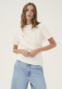 Top crema a maniche corte con collo alto e design drappeggiato. Abbinato a jeans azzurri chiari, dal taglio morbido. Texture liscia.