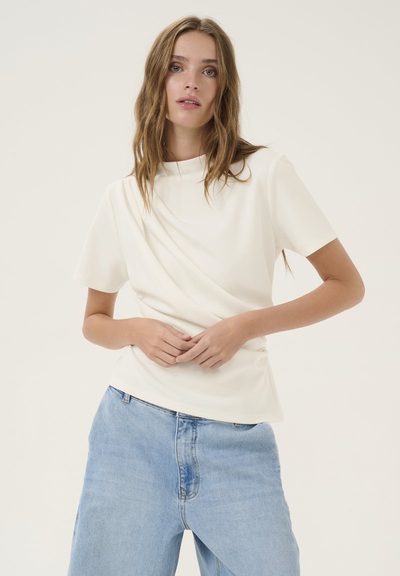 Top crema a maniche corte con collo alto e design drappeggiato. Abbinato a jeans azzurri chiari, dal taglio morbido. Texture liscia.