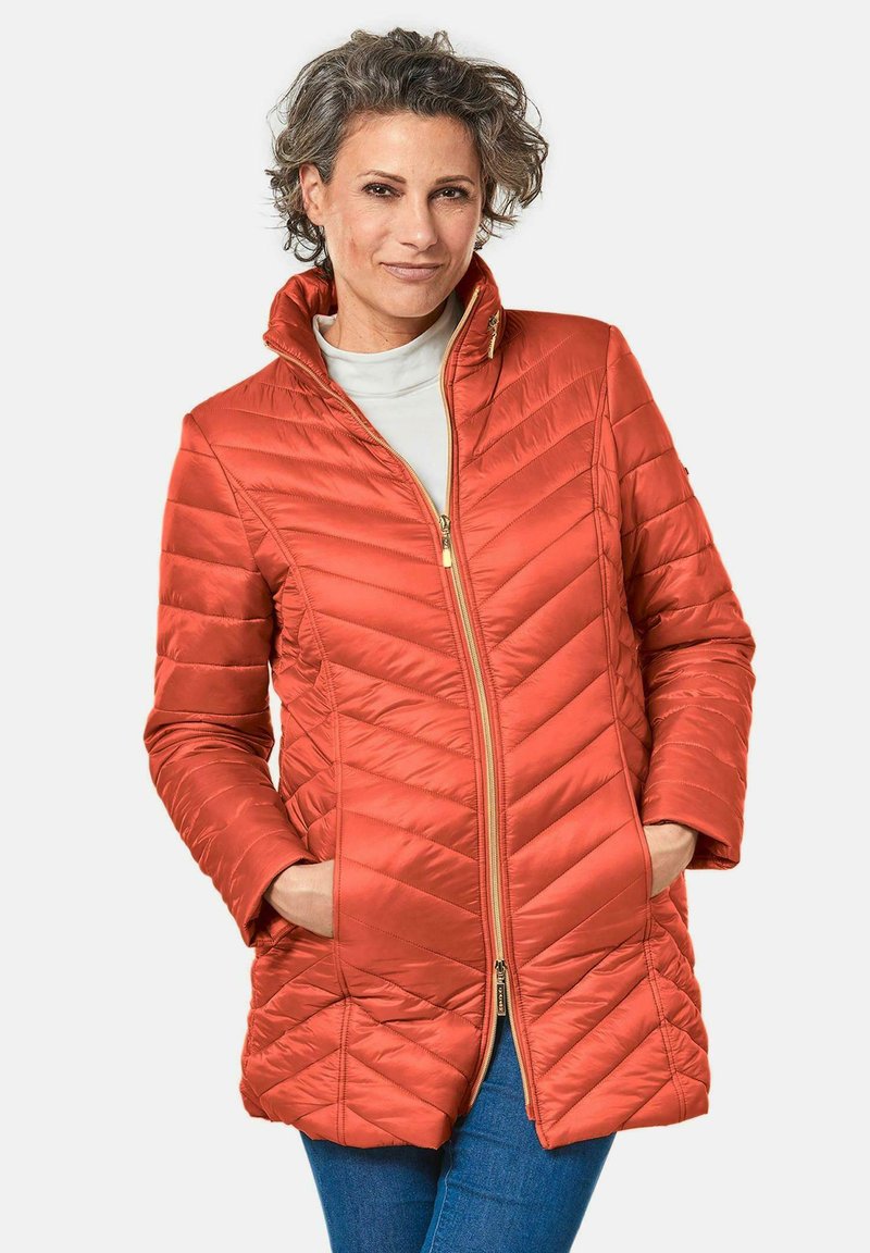 Gequilte, oranje synthetische jas met een hoge kraag, twee zijzakken en een gouden ritsopening. Kenmerkt zich door een chevron stikpatroon.