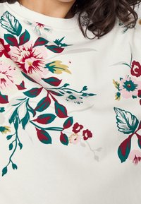 Vit sweatshirt med blommönster i nyanser av röd, rosa och grön. Har en rund halsringning och mjukt, texturerat tyg.