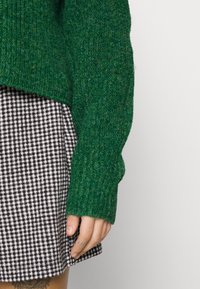Zielony sweter z dzianiny o teksturowanym wzorze, w połączeniu z czarno-białą spódnicą w kratę. Sweter ma długie rękawy i luźny krój.