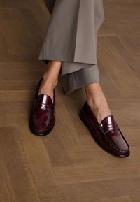 Burgunderfarbene Lederloafer mit genähten Details, runder Spitze und schwarzer Sohle, kombiniert mit hellgrauen, maßgeschneiderten Hosen. Hintergrund mit Holzfußboden.