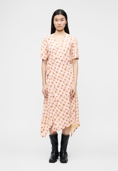 Femme portant une robe portefeuille rose à fleurs avec des manches courtes et un ourlet asymétrique, associée à des bottes en cuir noires montant jusqu'à mi-mollet.