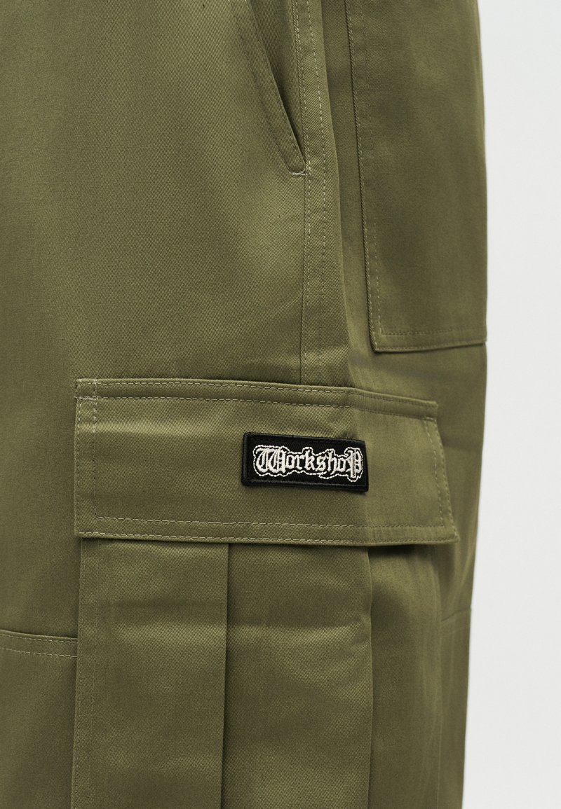Pantalones cargo verde oliva con solapa de bolsillo lateral etiquetada como "Workshop" en fuente gótica blanca sobre parche negro.