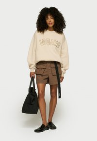 Sudadera beige con estampado "10DAYS" en marrón, combinada con pantalones cortos cargo marrones con bolsillos frontales y zapatos negros sin cordones. Incluye bolsa de mano negra.