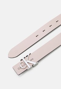 Cintura in pelle rosa chiaro con una fibbia in argento con logo "CK". Presenta fori uniformemente distanziati per la regolazione e una finitura liscia.