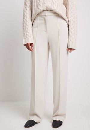 Persona que lleva pantalones anchos color crema con pliegues delanteros, combinados con un jersey beige de punto con trenzas y zapatos planos negros.
