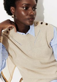 Pull en tricot beige avec des embellissements sur les épaules porté sur une chemise rayée bleu clair. Texture lisse, col rond et couture décorative.