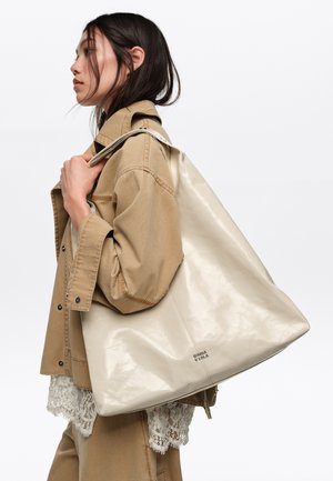 Joven mujer con cabello largo y oscuro lleva chaqueta y pantalones beige, llevando un gran bolso tote crema brillante con el nombre de la marca visible en el hombro.