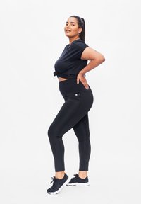 Zwarte sportoutfit bestaande uit een knoop-crop top, hoge taille leggings en zwarte hardloopschoenen met witte zolen, gepresenteerd op een effen achtergrond.