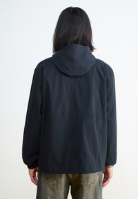Jachetă cu glugă neagră, cu o croială lejeră, realizată dintr-un material texturat și cu manșete elastice. Purtați-o peste o pereche de pantaloni din denim albastru deschis.