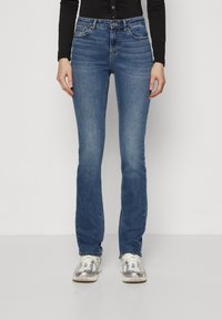 Kék farmernadrág, slim fit szabású, enyhe harangszárral a bokánál. Öt zsebbel és kontrasztos varrással rendelkezik. Sportos cipővel párosítva.