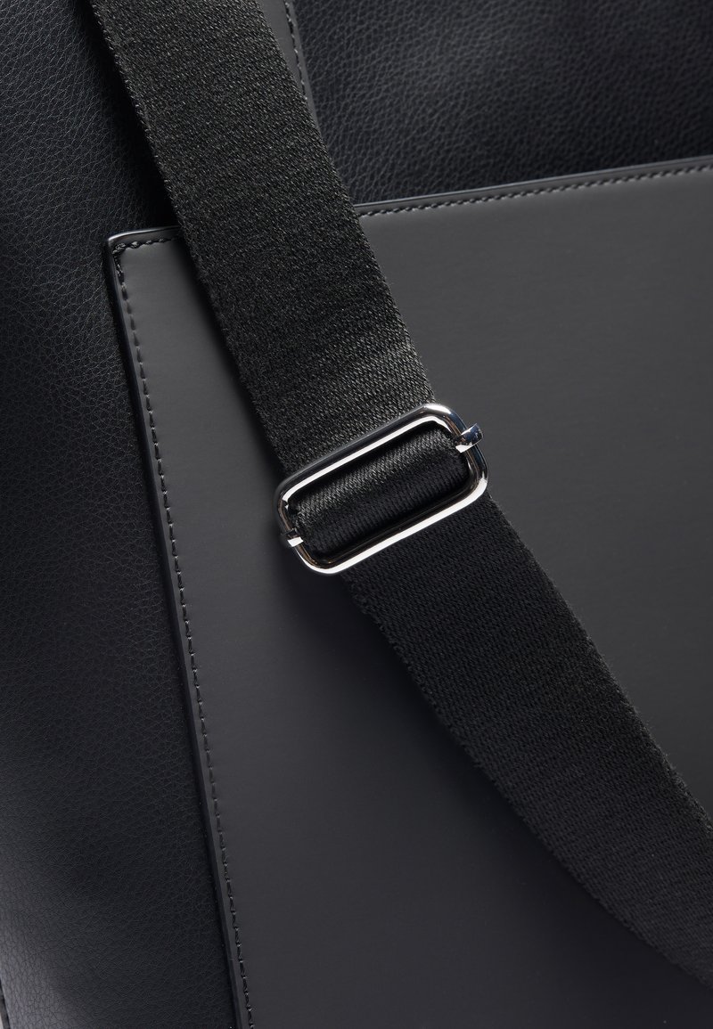 Sac en cuir noir avec une texture lisse, comportant un panneau plat, une sangle en tissu noir et une boucle métallique argentée pour le réglage.