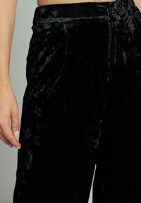 Zwarte fluwelen broek met een gladde textuur, voorzien van een hoge tailleband, zijzakken en een lichte glans. Close-up van een hand op de stof.