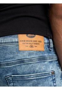 Blue denim jeans with a faded wash, featuring a leather label that reads "Tetrov" and details about crafting quality garments.