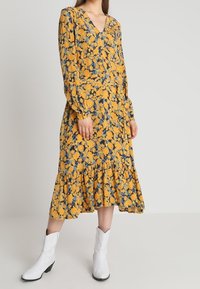 Vestido floral amarillo con acentos azules, escote en V, mangas largas y dobladillo con volantes. Combinado con botas blancas hasta el tobillo.