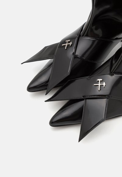 Stivaletti alla caviglia in pelle verniciata nera con punta appuntita e cinturini incrociati decorati con accenti metallici. Finitura liscia e lucida.