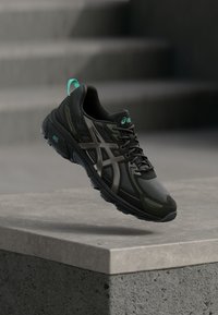 Shoe atlético preto com parte superior de malha, detalhes em prata e marcação em teal. Possui sola acolchoada e aderência texturizada para desempenho.