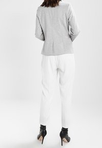 Šedý přiléhavý blazer s hladkým povrchem v kombinaci s bílými kalhotami s proužky. Kalhoty se u kotníků zužují a jsou doplněny černými botami na podpatku.