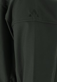Veste extérieure vert foncé en tissu léger, avec poches zippées, coutures plates et un logo subtil en broderie contrastée.