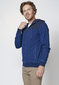 Blue zip-up hoodie made of soft fabric with ribbed texture on shoulders, front logo detail, and drawstring hood. Features side pockets.