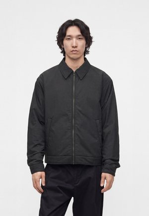 Jeune homme aux cheveux mi-longs foncés, portant une veste noire zippée et un pantalon noir, debout devant un fond clair uni.