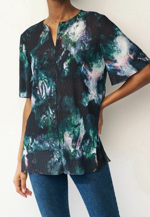 Vrouw draagt een kortemouw, geribde, ritssluiting voor blouse met een donker groen, wit en paars abstract patroon, gecombineerd met blauwe spijkerbroek.