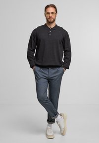 Mann mit hellbraunen Haaren und Brille steht mit den Händen in den Taschen, trägt ein dunkles Langarmpolo, blaue Hosen, weiße Socken und Turnschuhe.