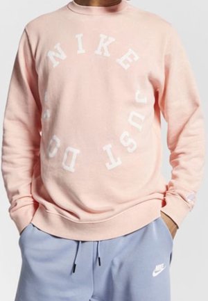 Osoba nosząca jasnoróżową bluzę Nike z napisem „NIKE JUST DO IT” ułożonym w okrągły wzór, w połączeniu z jasnoniebieskimi spodniami dresowymi Nike.
