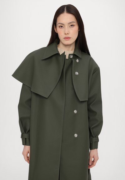 Πράσινο αδιάβροχο παλτό με έναν χαρακτηριστικό γιακά τύπου κάπας, με ασημένια κουμπιά και μακριά μανίκια με ρυθμιζόμενους καρπούς. Λείος υφάσματος.