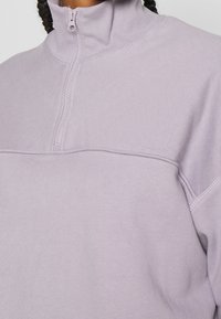 Světle fialový pulovr na čtyřčtvrťový zip vyrobený z měkké látky. Má vysoký límec a prošitou šev přes hrudník pro přidaný detail.