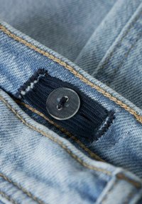 Gaiši zils džinsa audums ar redzamu šūtu šuvi un brūnu diegu. Skatāms caurspīdīgs plastmasas pogas stiprinājums pie tumši zila auduma cilpas.