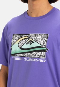 Camiseta de algodón morada con un logo gráfico que presenta un diseño de montaña en verde y amarillo sobre un fondo negro moteado.