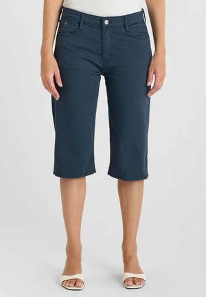 Vrouw draagt marineblauwe knielange shorts en witte open-toe hakken, staand tegen een effen lichte achtergrond.