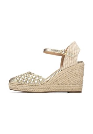 Sandalia espadrille de cuña con tacón de yute tejido, parte delantera metálica dorada tejida y tira en el tobillo con hebilla dorada sobre fondo blanco.