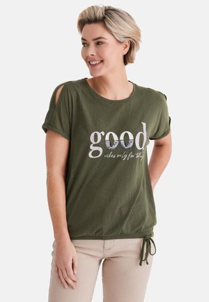 Lächelnde Frau mit kurzen blonden Haaren, die ein olivgrünes Hemd mit hochgekrempelten Ärmeln und dem Schriftzug „good vibes only for today“ sowie beigefarbene Hosen trägt.