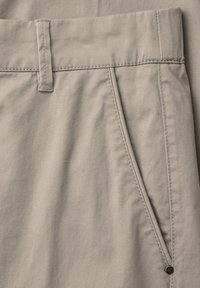 Beige stof in close-up met zicht op tailleband, riemlus, zijzak met metalen klinknagel en zichtbare stiksels op de broek.