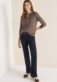 Brązowa bluzka z długim rękawem, dekoltem w serek i przeszyciami, zestawiona z ciemnoniebieskimi dzwonami oraz brązowymi trampkami z białymi akcentami.