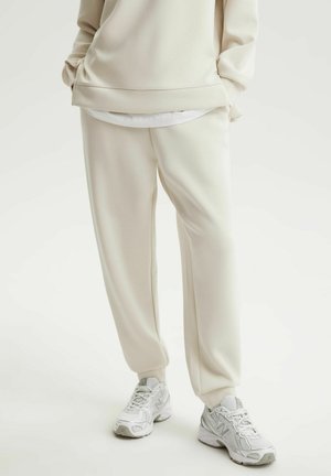 Dolna część ciała ubrana w kremowe spodnie dresowe, pasującą bluzę, białą podkoszulkę i białe buty New Balance, stojąca na jednolitym białym tle.