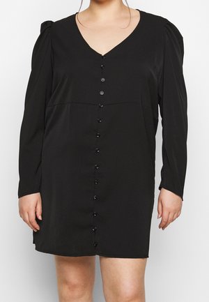 Kvinde iført en sort mini-kjole med lange ærmer, forreste knapper og V-hals mod en ensfarvet baggrund.