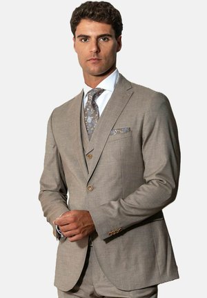 Man wearing beige three-piece suit with floral patterned tie and pocket square, standing against plain light background.