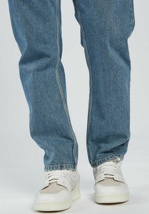Helles blaues Denim-Jeans mit lockerer Passform, sichtbar genähten Nähten und einem leicht fransigen Saum, kombiniert mit weißen Sneakern.