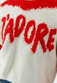 Gebreide trui met een crème basis, een pluizige rode tekst "J'ADORE", gestructureerd garen en een relaxte pasvorm.