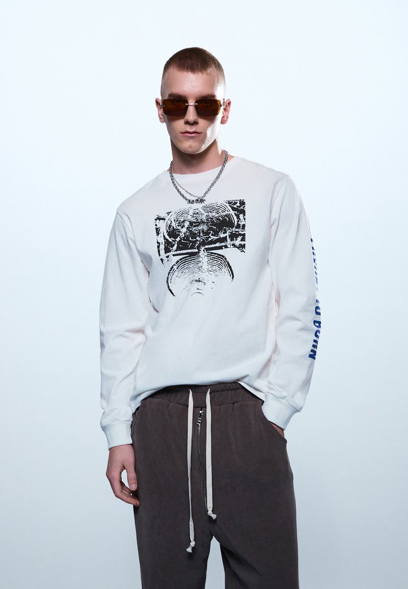 Vyras trumpais plaukais, dėvintis rudus saulės akinius, baltą ilgomis rankovėmis su grafiniu raštu marškinį, sidabrinį grandinėlės vėrinį ir tamsiai rudus kelnės su raišteliais.