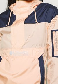 Lätt anorak i persika- och marinblå färg, med en halvdragkedja, justerbar huva och ficka med dragkedja och nät detaljer.